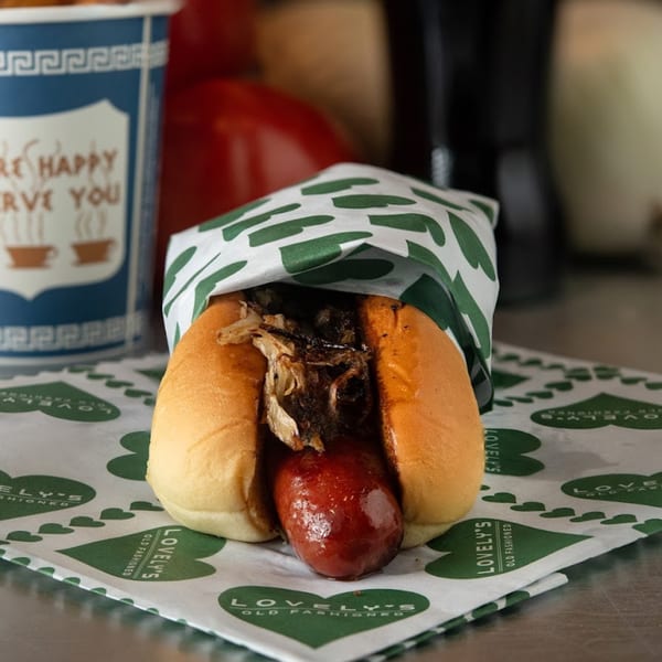 A hot dog with caramelized onions in a bun, wrapped in Lovely's Old Fashioned paper.