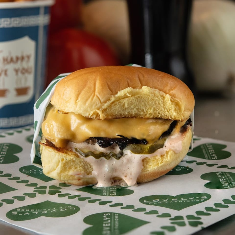 A Lovely's Old Fashioned burger with melted cheese, pickles, and sauce on branded paper.