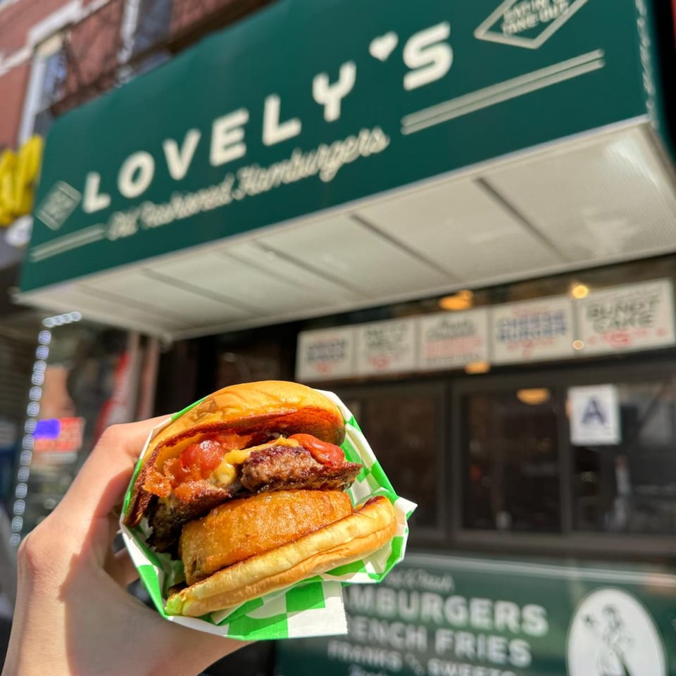 A hand holds a burger from Lovely's Old Fashioned, with the restaurant's green awning in the background.